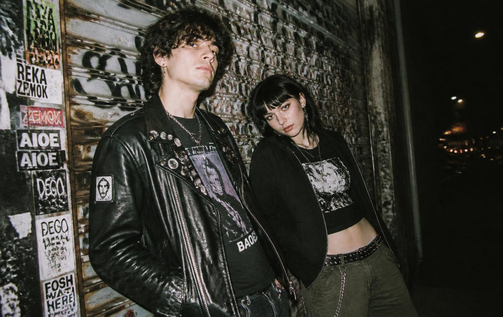 Two models in punk streetwear against a sticker-covered shutter at night